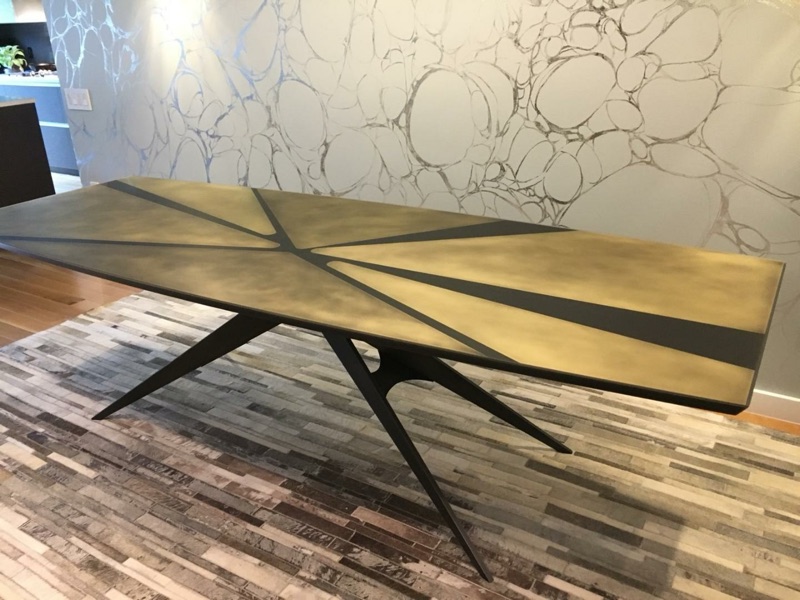 Statement dining table with a brass-toned resin surface on a blackened-steel starburst base, set against a marble wall, by Van Aken Design