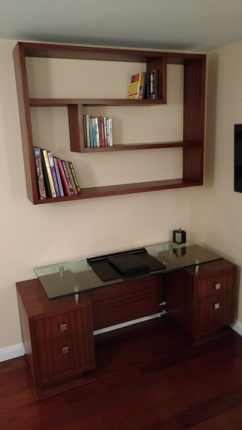 Custom home office desk in warm hardwood with a glass top and a floating geometric wall shelving unit by Van Aken Design